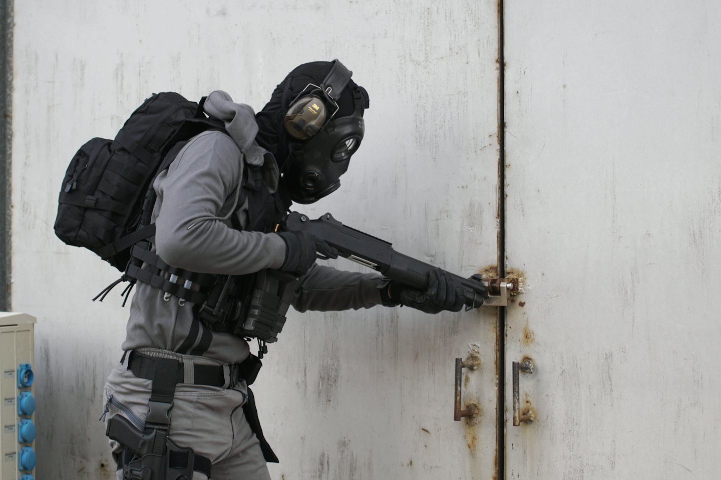 Person in tactical gear opening a rusted padlock with a shotgun.